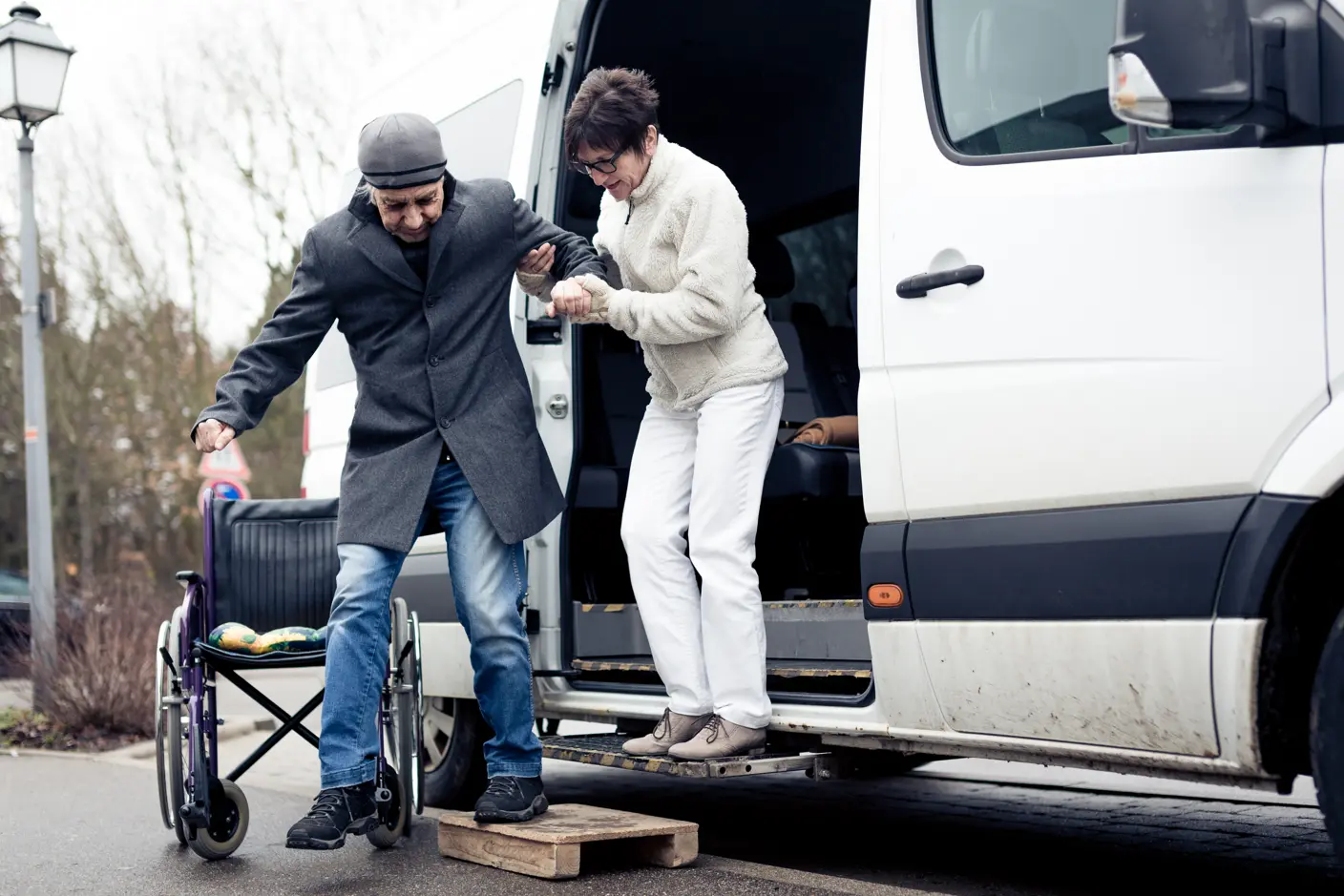 A clinician helping a patient out of a vehicle into a wheelchar, practicing safe transport best practices for preventative care. Both are wearing winter jackets